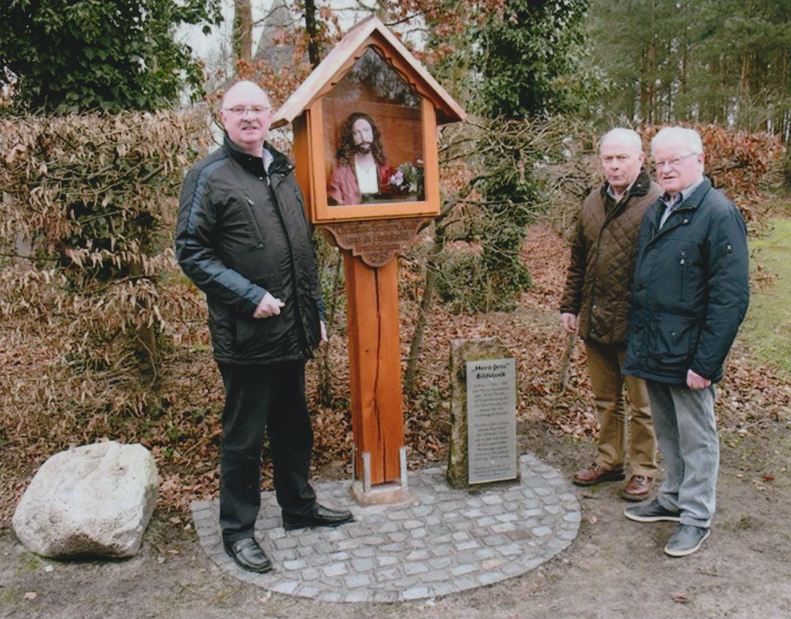 Drei Männer stehen neben dem renovierten Herz-Jesu-Bildstock, davor ein Pflasterkreis und die Dokumentationstafel