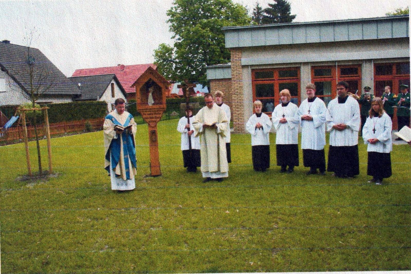 Geistliche und Messdiener stehen vor dem Bildstock auf einer Wiese, dahinter der Kindergarten
