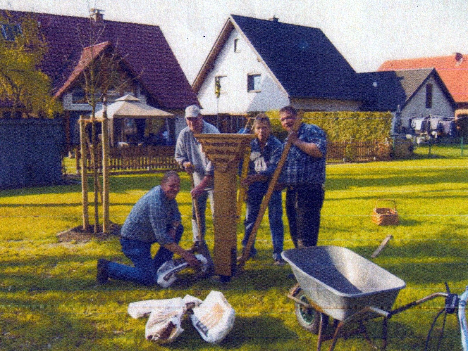 Helfer beim Aufstellen des restaurierten Bildstocks in einer Grünanlage, im Vordergrund eine Schubkarre