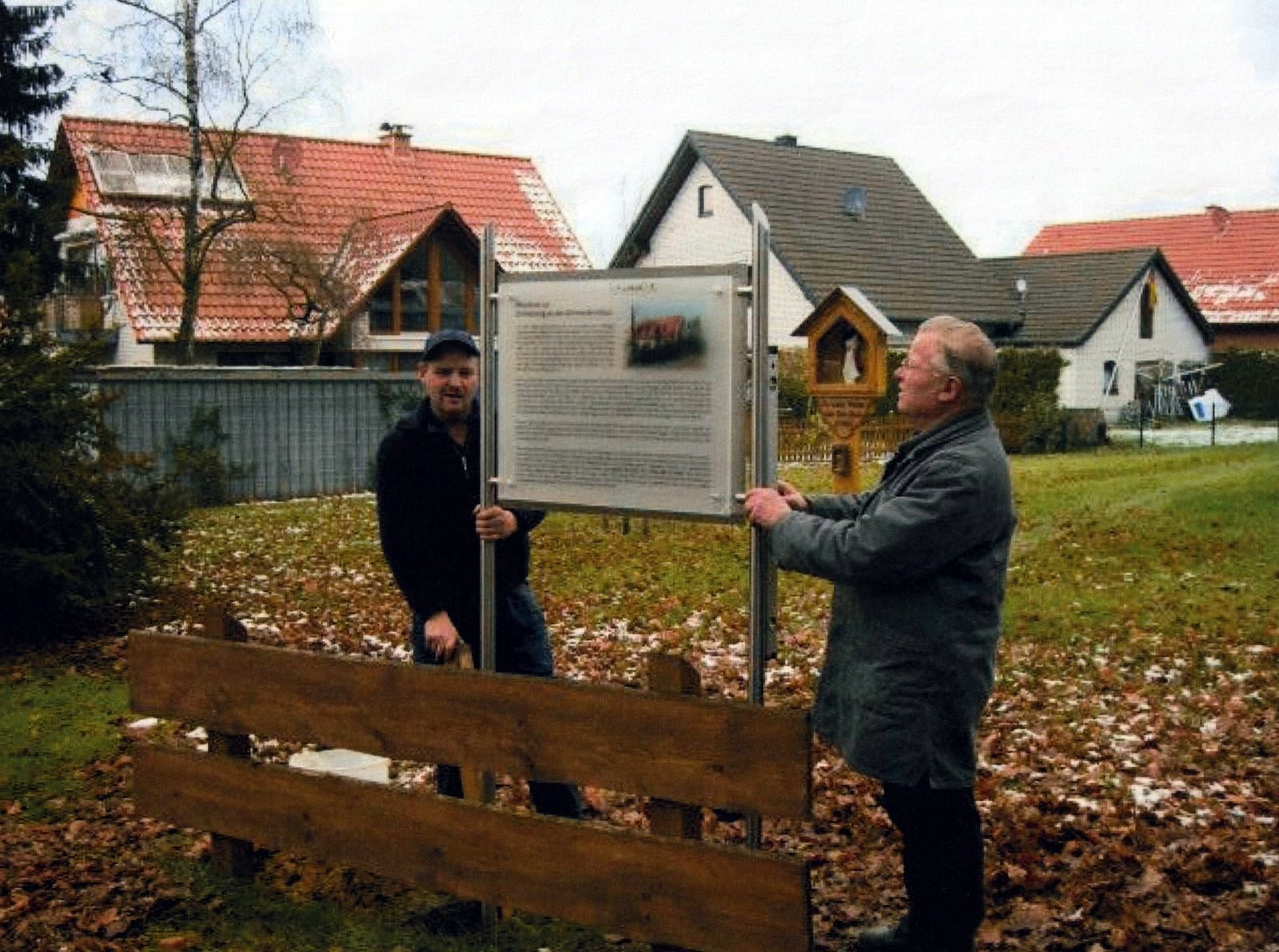 Zwei Männer halten die Gedenktafel auf ihrem Gestell, im Hintergrund der Bildstock und Wohnhäuser